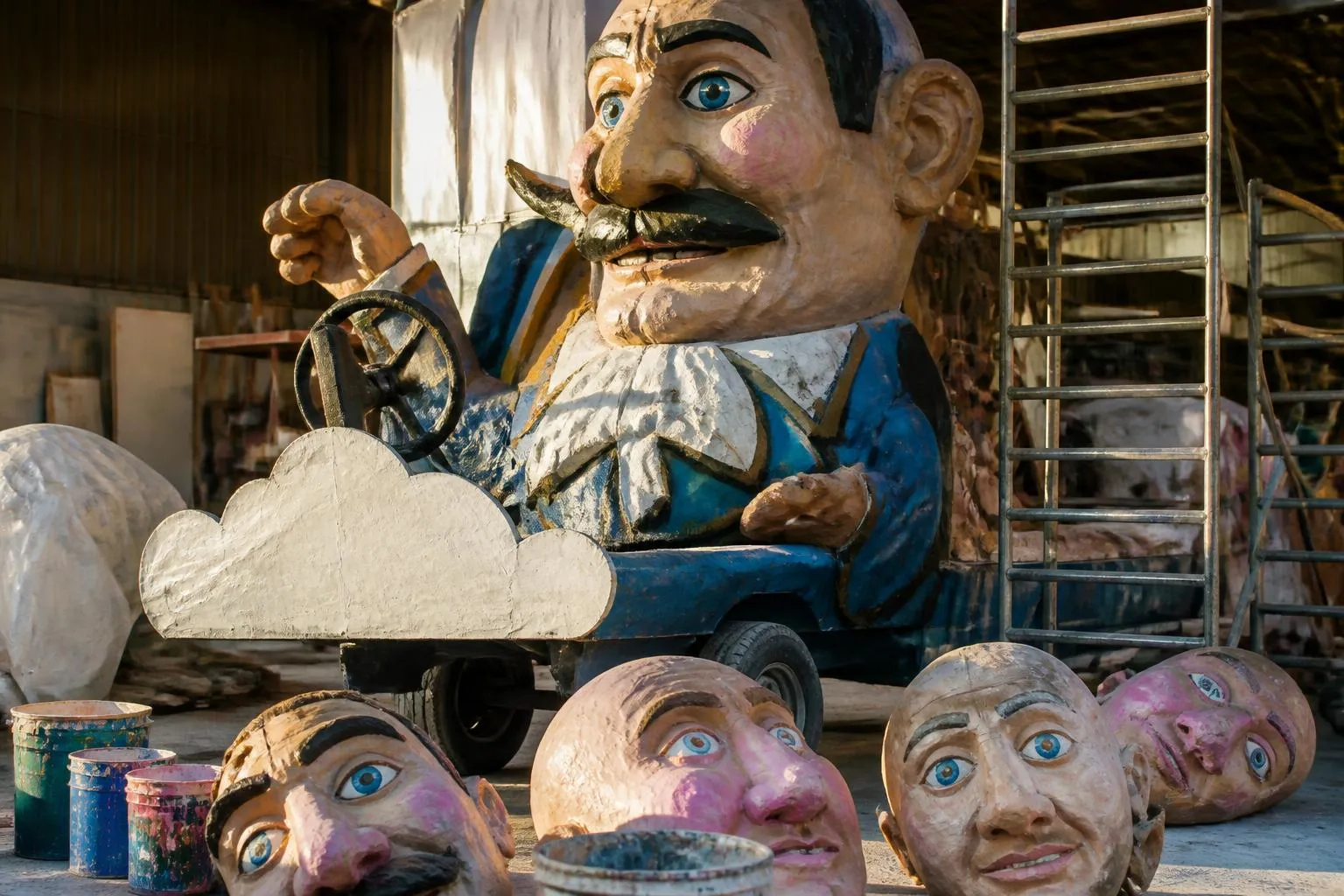Un char de carnaval en carton peint garé dans un hangar, entouré de grosses têtes en cours de finition